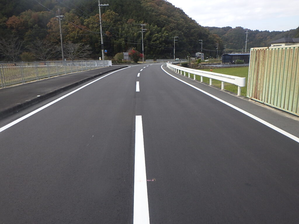 Two-lane rural road with a solid white centerline curving to the left, flanked by guardrails and a fence, hills in the distance.