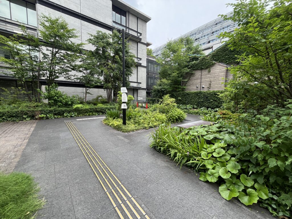 Modern urban courtyard with a gray brick building, lush trees, and a paved path featuring yellow tactile guiding strips for accessibility.