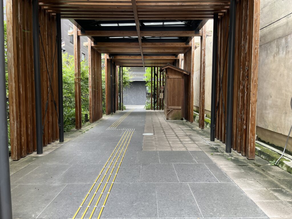 Wooden pillar walkway under a timber-framed cover, with yellow tactile guide lines along the path toward a small structure in the distance.