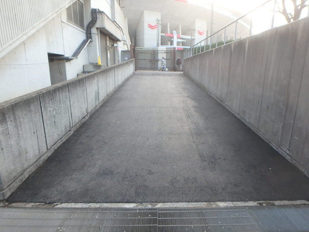 Concrete alley with high concrete walls, metal gate at far end, and a drainage grate in the foreground; two workers near the gate.