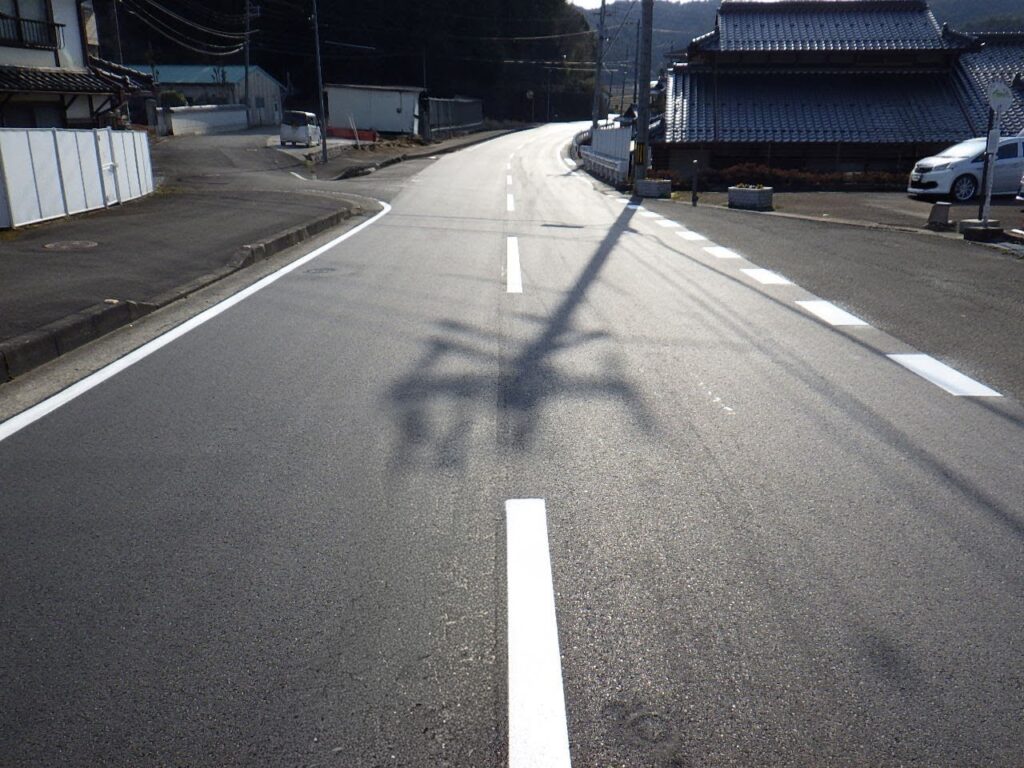 Residential street in Japan with white road markings and shadows from power lines; traditional tiled houses to the right.