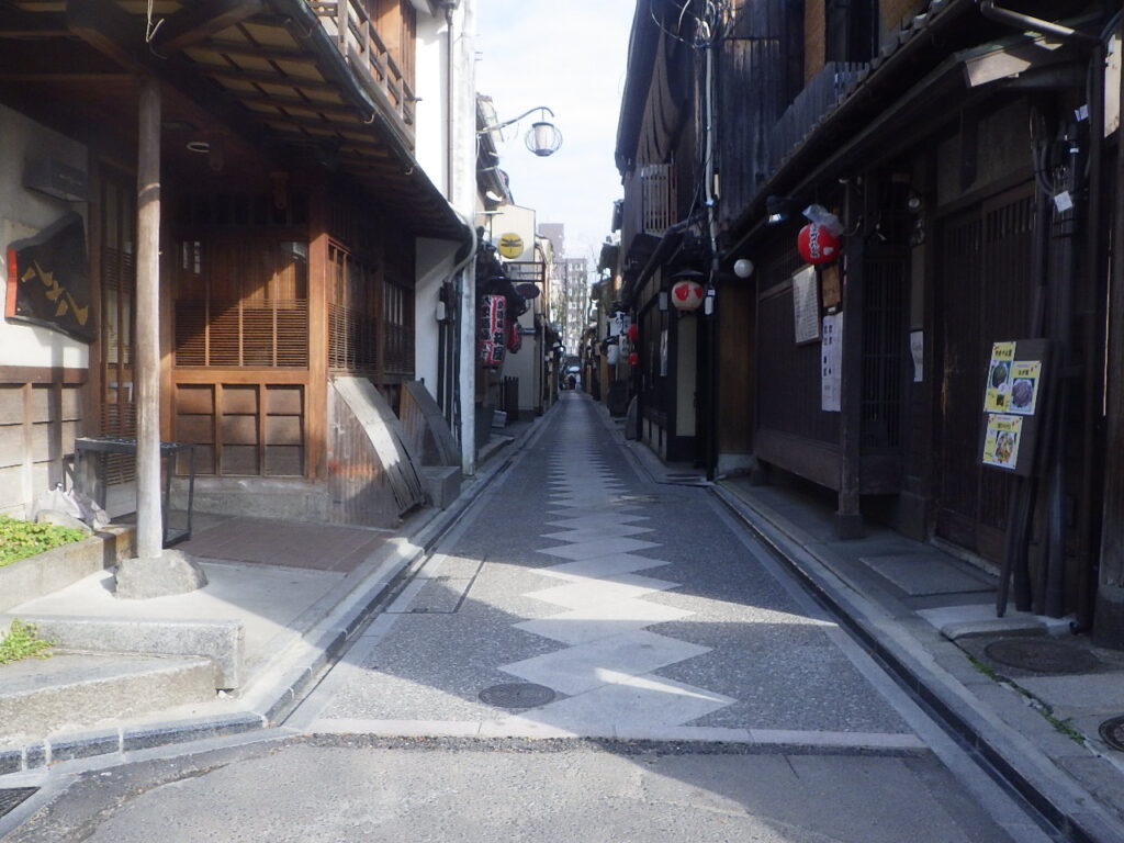 Narrow Japanese alley with wooden shopfronts, hanging lanterns, and a zigzag pedestrian paving pattern down the center street on a sunny day.
