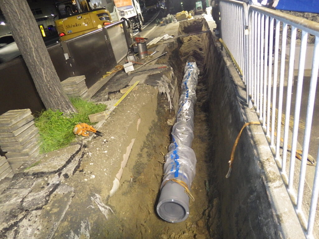 Construction trench along a sidewalk with a long wrapped pipe laid in the ditch and construction tools nearby at night.