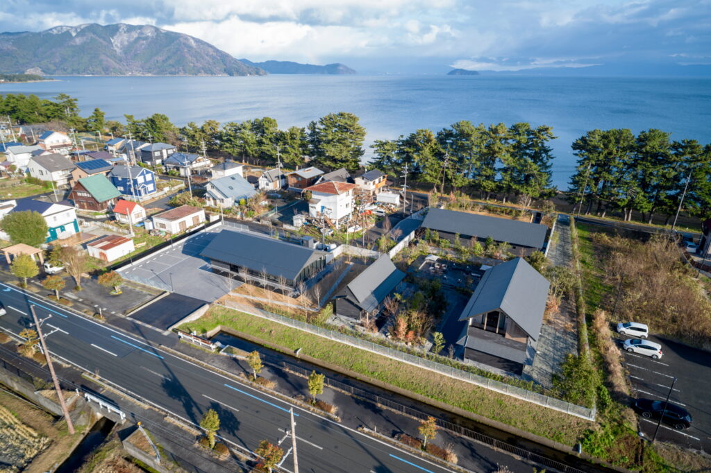 Aerial view of a coastal town with colorful houses along a grid-like street, row of trees by the shore, and calm blue sea with mountains in the distance.