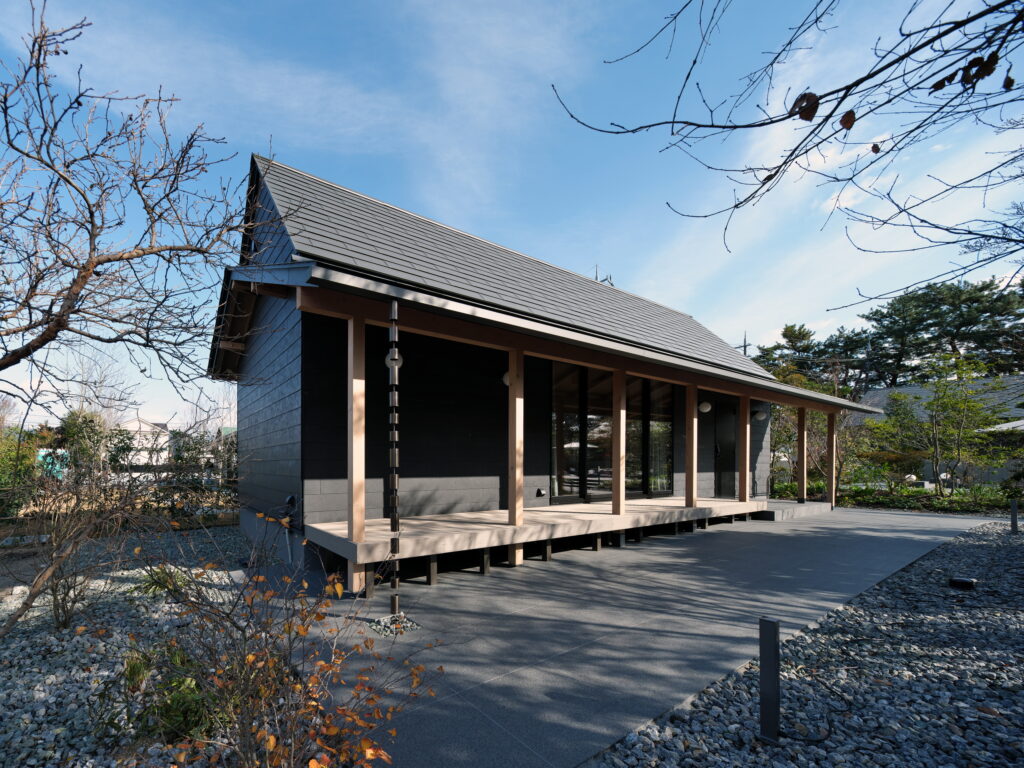 Modern dark house with a sloped metal roof and large glass doors, raised deck, leafless trees, blue sky.