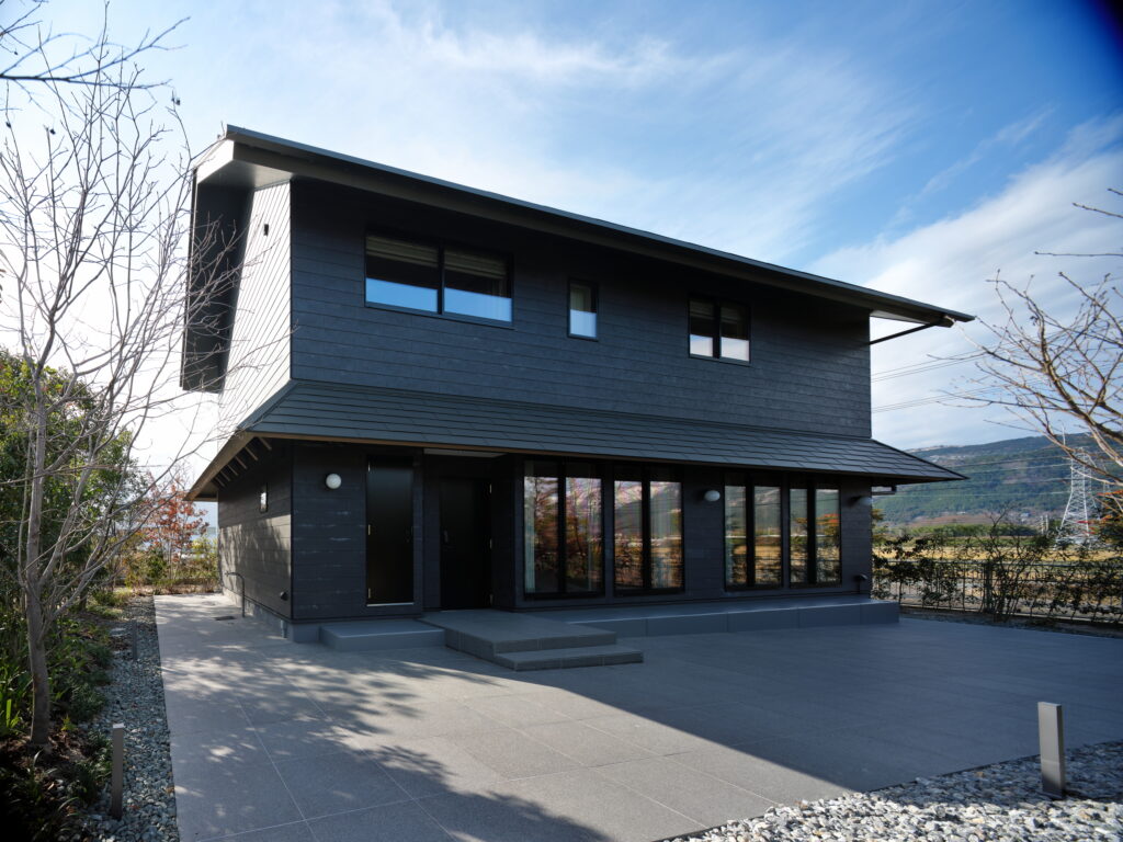 Modern dark gray two-story house with large glass windows and a slanted roof, a concrete front path, and sparse trees against a blue sky.