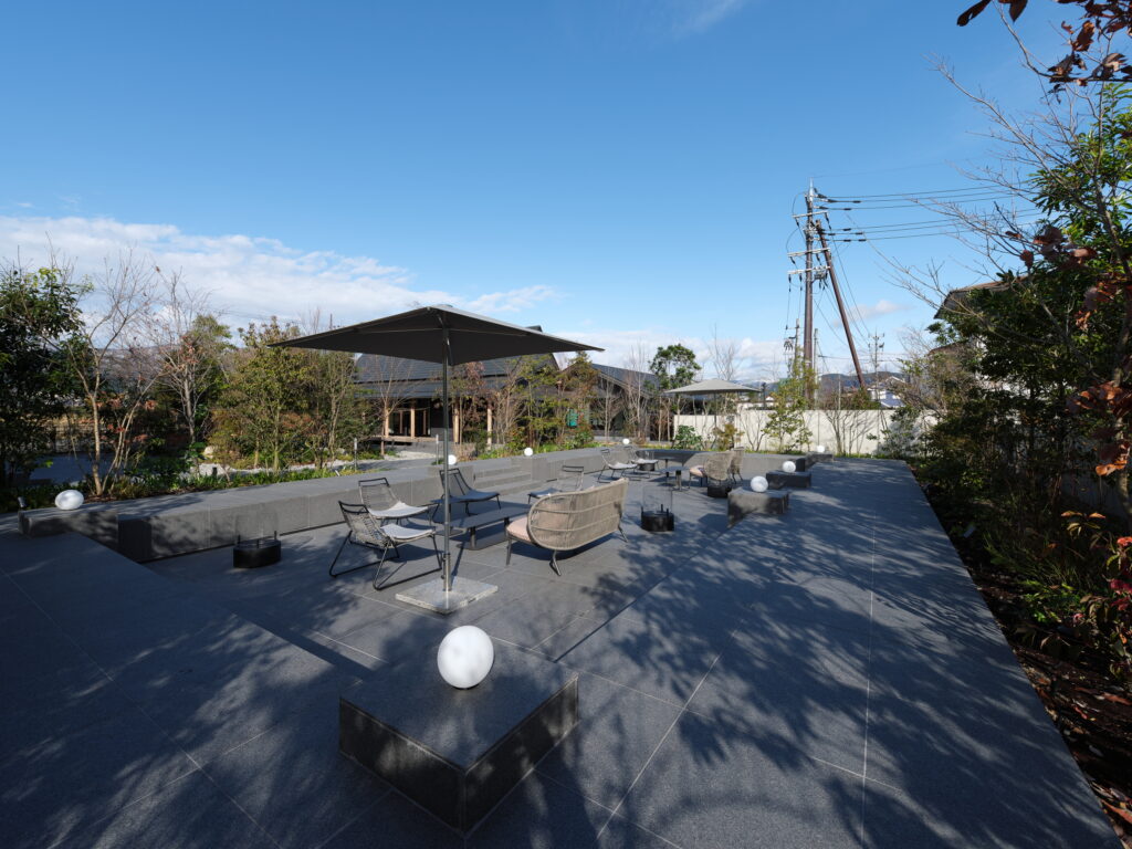Outdoor courtyard with gray tile flooring, lounge chairs, and large shade umbrellas under a clear blue sky.