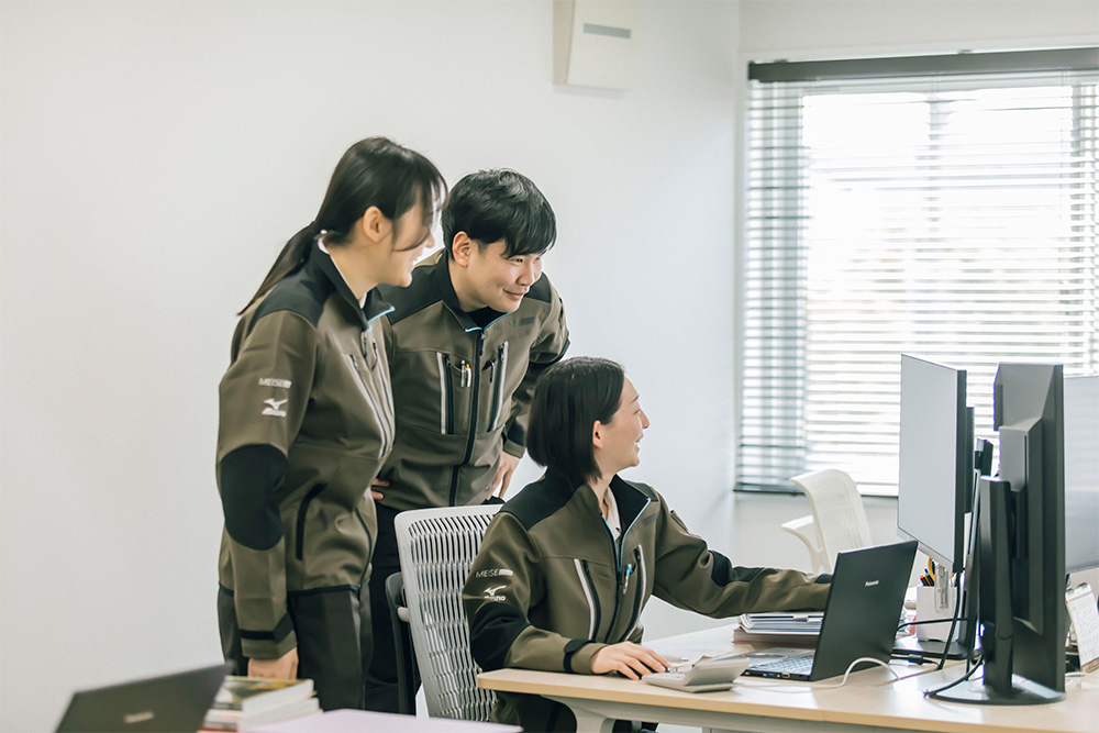 Three coworkers in olive uniforms gather around a desk, looking at a computer screen with dual monitors.