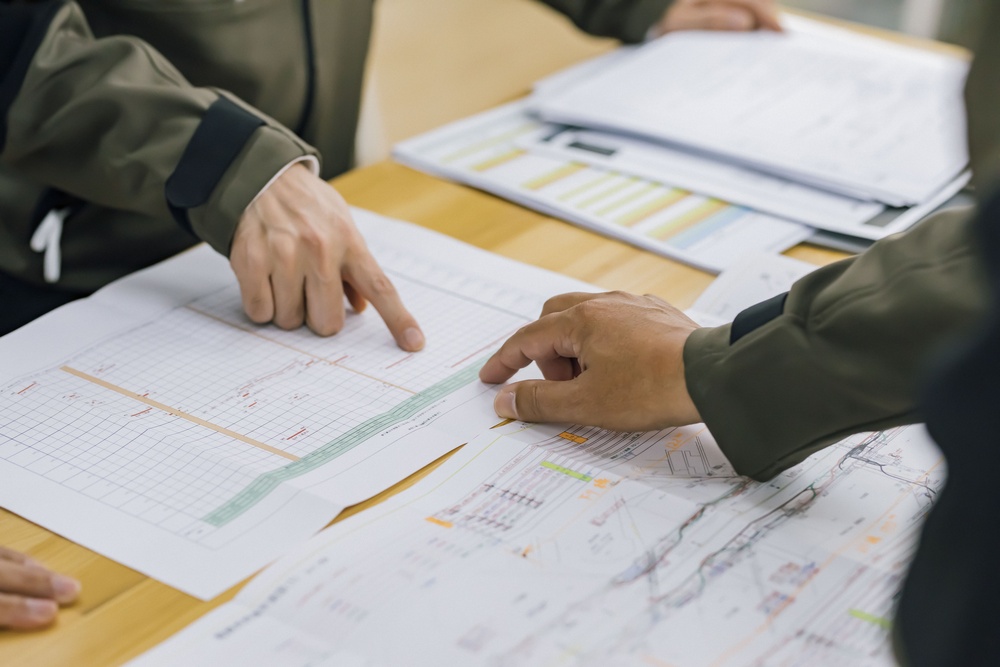 Two people study architectural blueprints on a wooden table, with one pointing at details.