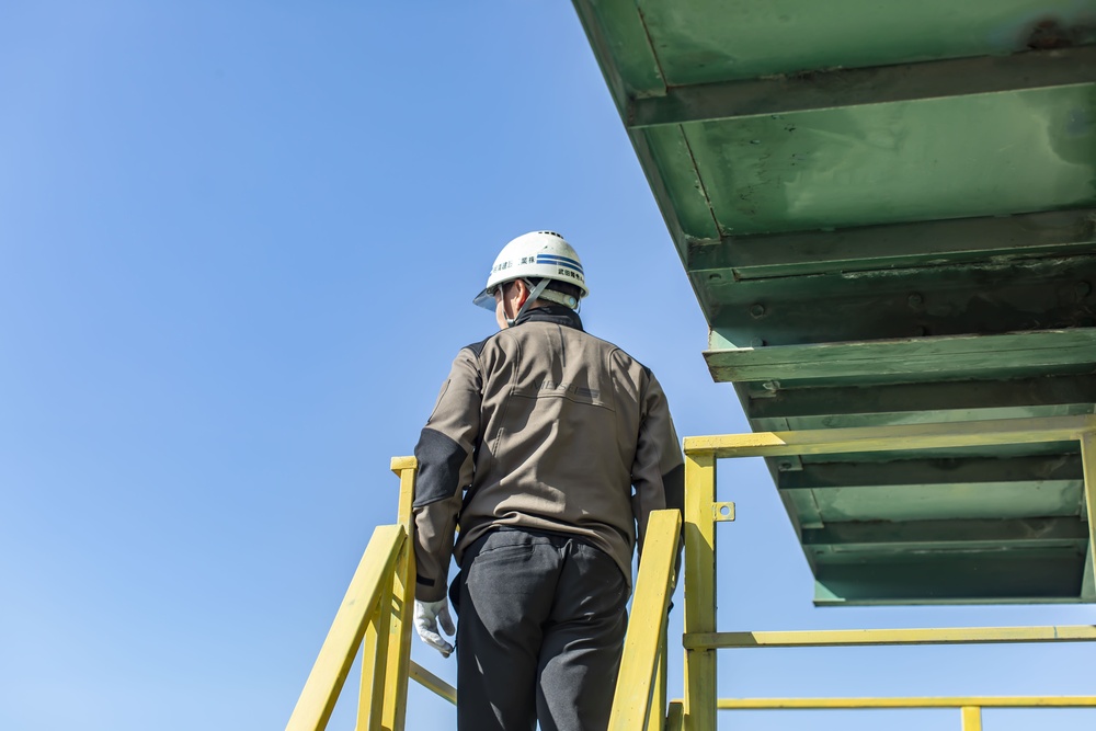 Back view of a construction worker in a hard hat and brown jacket standing on yellow stairs against a clear blue sky.