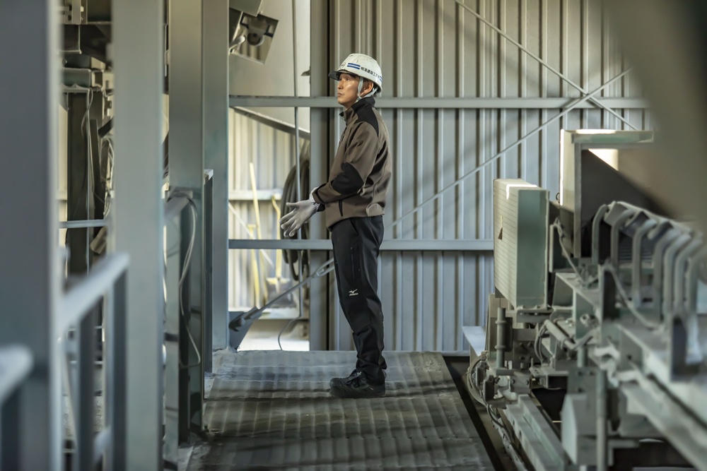Worker in safety helmet and jacket inspecting industrial equipment on a catwalk area.