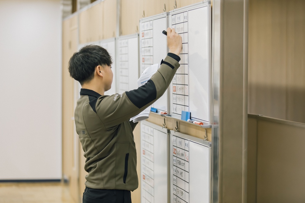 Young man in a green jacket writes on a wall-mounted whiteboard filled with calendars and notes in a bright room, updating schedules.