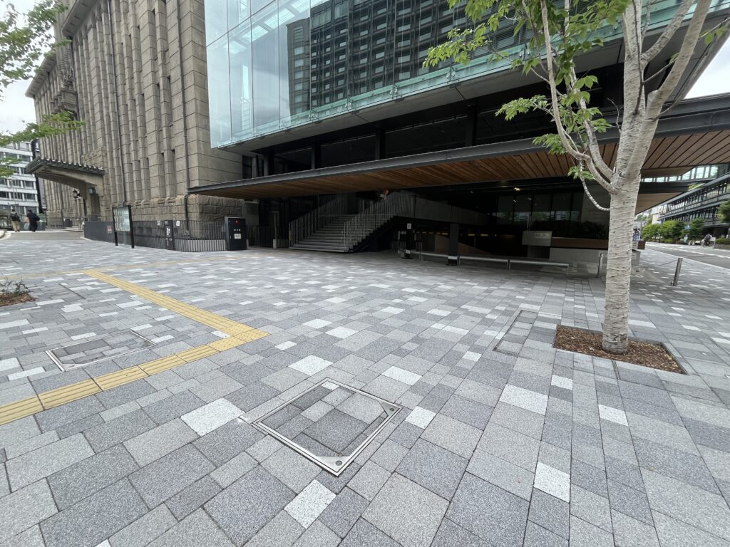 Wide view of a modern urban plaza with gray tiled ground, a large stone building, and a tree in the foreground on the right.
