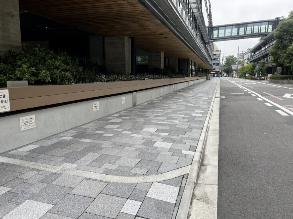 Wide sidewalk with gray patterned pavers beside a modern building and green planters; street to the right and a distant pedestrian bridge.