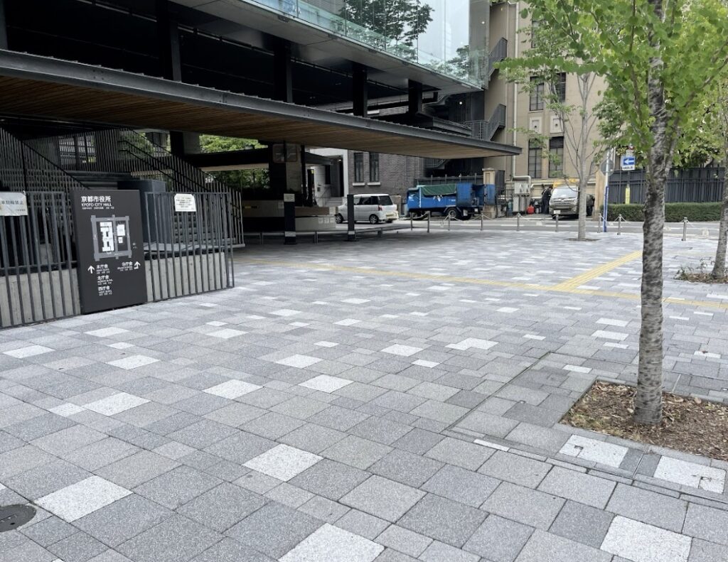 Wide view of a modern building entrance with a covered drop-off area, a metal fence and sign, and parked white van and blue truck in the background on a tiled plaza side road.