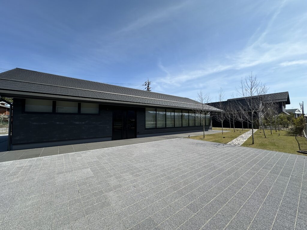 Modern dark-grey, single-story building with large windows, a paved plaza, and leafless trees against a blue sky.