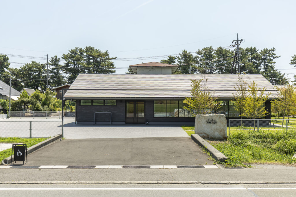 Modern, single-story dark building with large front windows and a tan sloped roof, in a paved lot with a stone sign reading 'Tala Voyage' in the foreground.