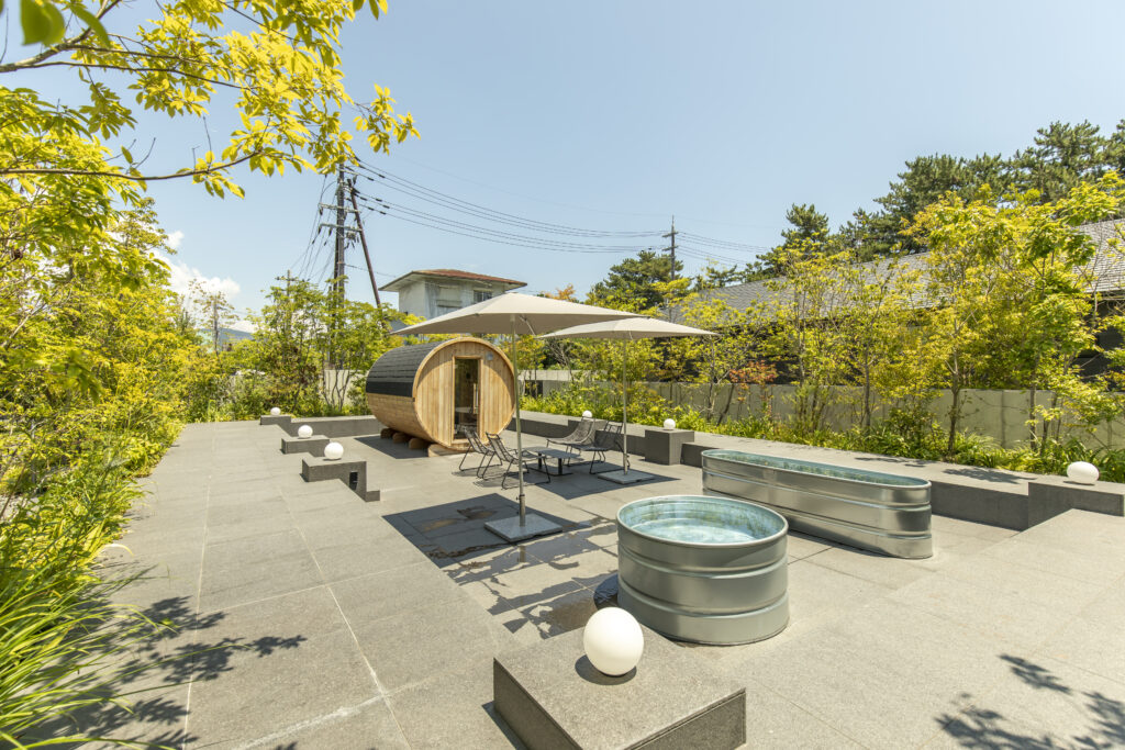 Outdoor patio scene with a round wooden barrel sauna, white umbrellas, and metal chairs amid greenery under a blue sky, bright and serene.