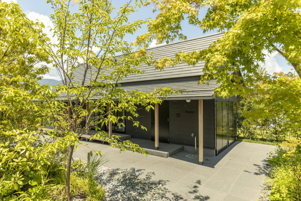 Modern dark-gray building with a wooden entrance canopy, framed by lush green trees on a sunny day