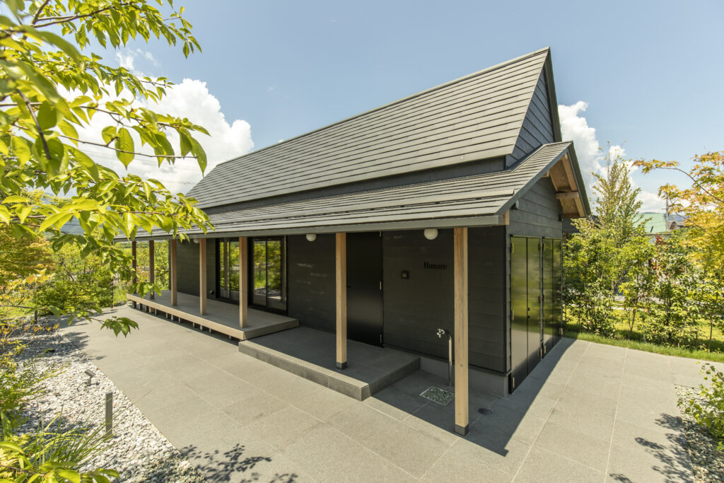Modern dark-gray house with a large sloped metal roof, wooden posts, and glass doors, set among greenery against a blue sky.