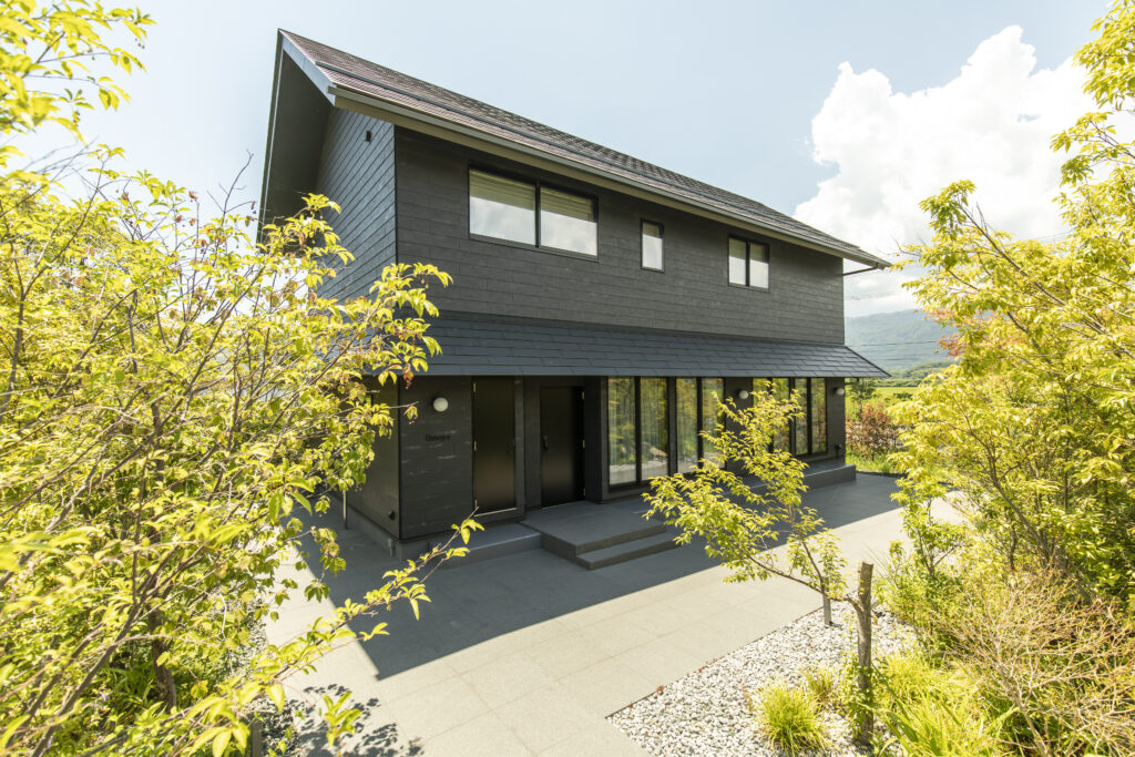 Modern dark gray two-story house with large windows, surrounded by green trees on a sunny day of bright sky.